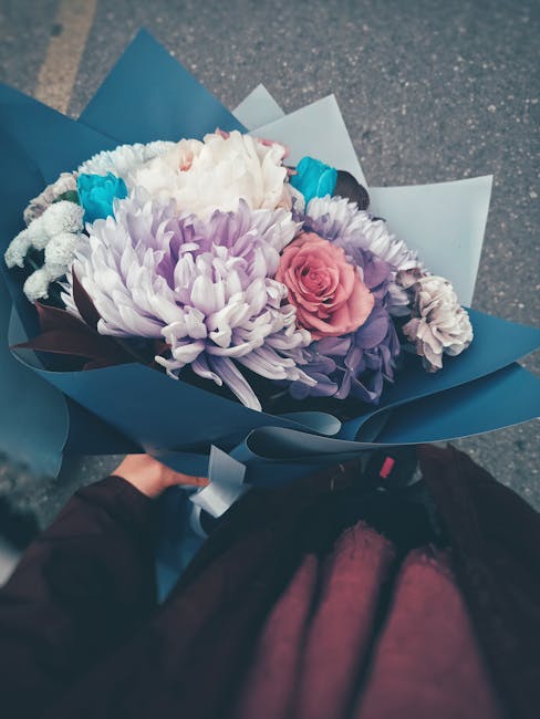 A hand holds a bouquet of fresh flowers wrapped in blue and white paper. The arrangement includes a large white chrysanthemum, soft pink roses, light purple lisianthus, and blue accent flowers, all wi
