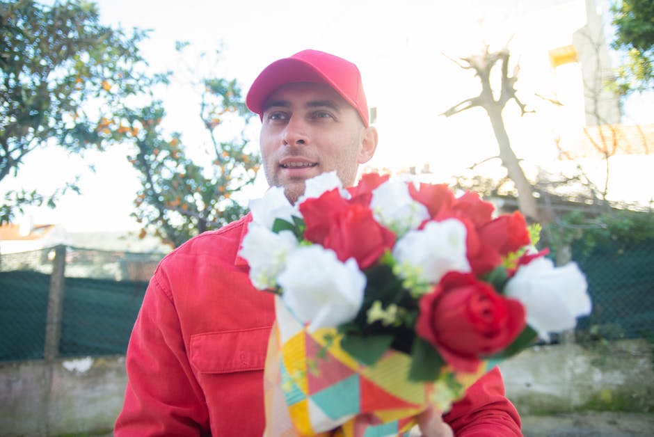 A delivery person dressed in a red uniform and cap is holding a bouquet of fresh flowers in front of him. The bouquet includes red and white roses, with some green foliage visible, wrapped in colorful