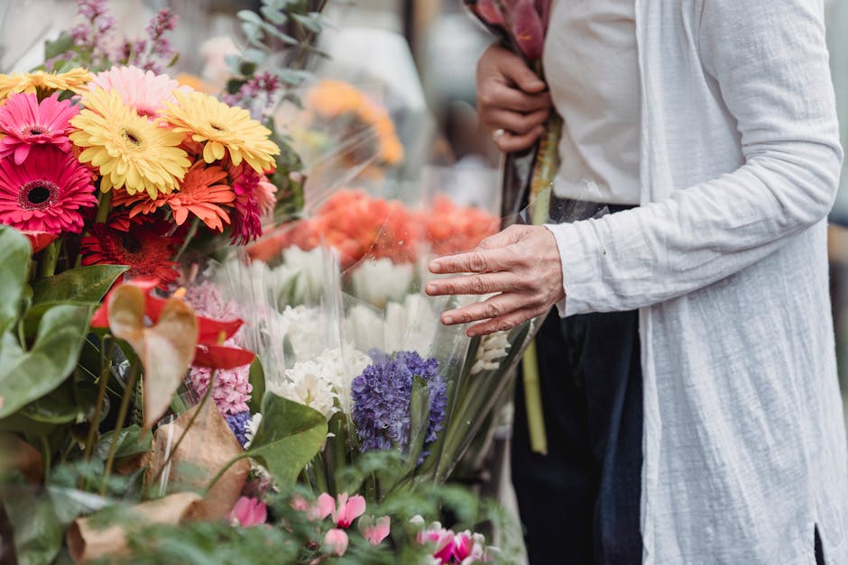 A person dressed in a light grey cardigan and dark trousers is browsing a vibrant flower stall, examining a bouquet wrapped in transparent plastic. The display features a colorful selection of fresh f