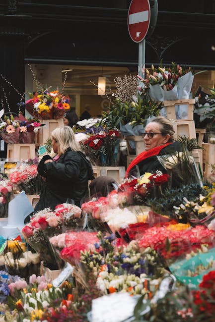 A busy floral market scene with an assortment of fresh flower bouquets and arrangements displayed on wooden shelves and tables. The flowers include bright pink, white, red, yellow, and purple blooms,