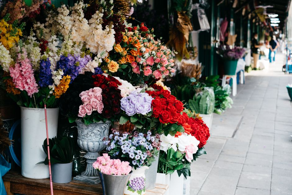 A vibrant outdoor flower stall displays an array of fresh floral arrangements in various containers, including white buckets and ornate vases, featuring a diverse selection of roses in shades of pink,