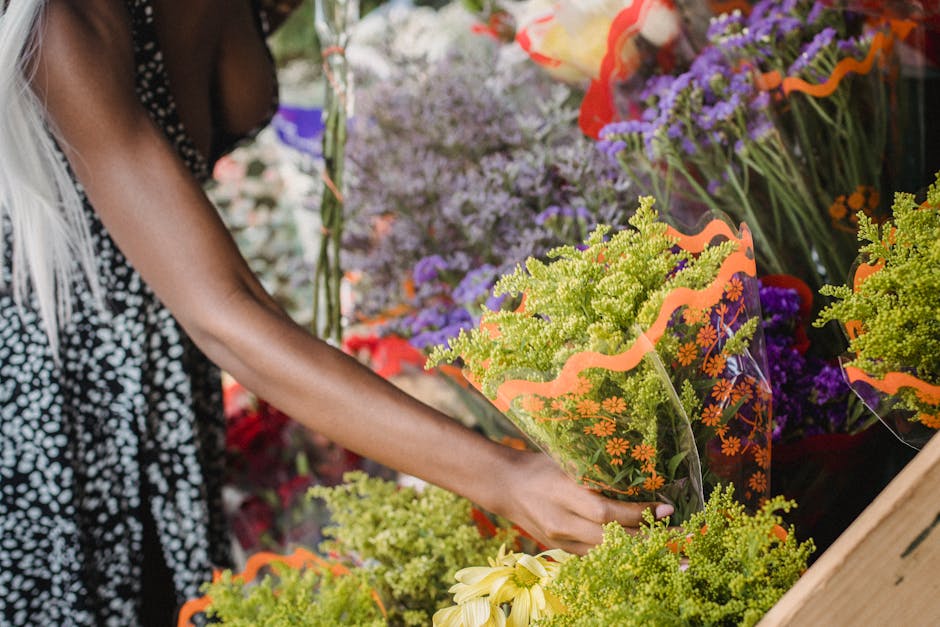 A person browsing a floral display at a flower shop, selecting a bouquet wrapped in clear plastic with orange floral patterns. The bouquet contains bright green foliage and small yellow flowers, and i